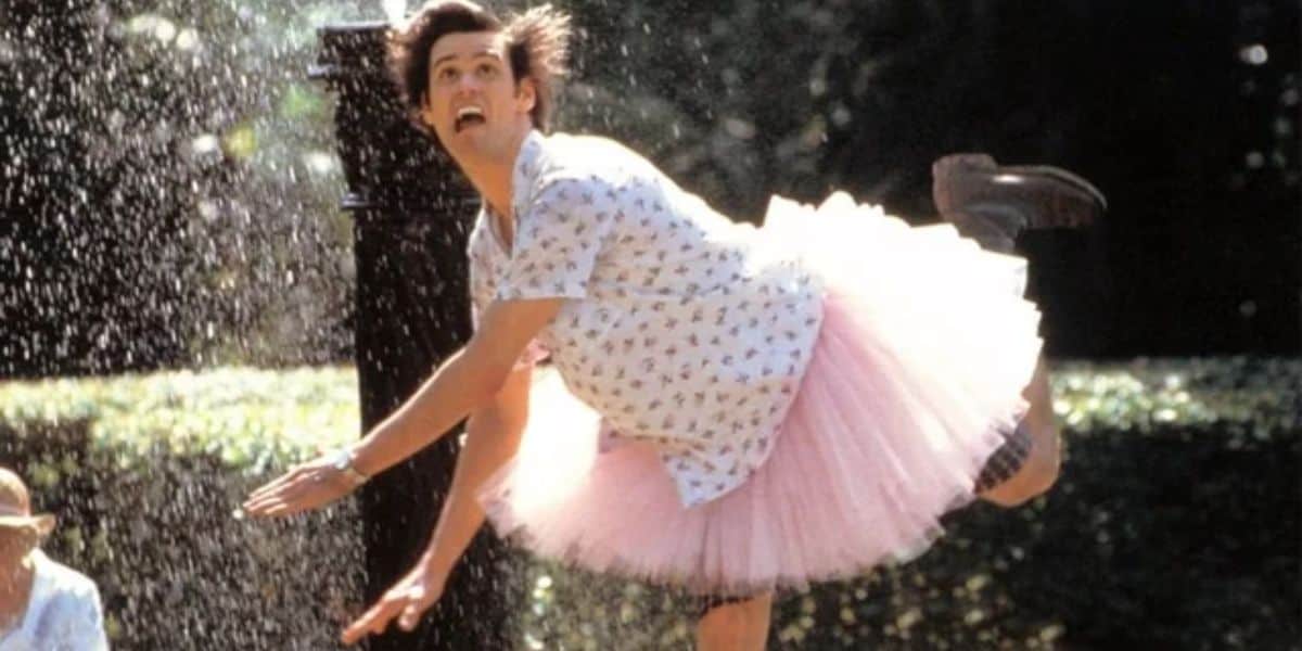 Ace Ventura (Jim Carrey) in a pink tutu leaping joyfully through a sprinkler on a sunny day. 