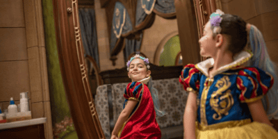 Two young girls in costume play backstage; one dressed as snow white and the other in a red and blue outfit, both smiling in a room with mirrors and stage props.