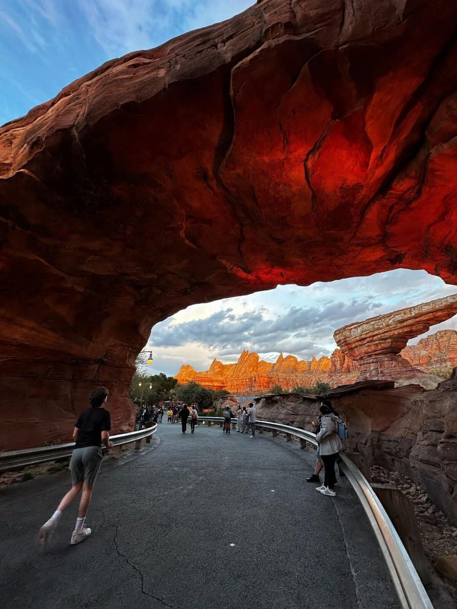 A rock tunnel in Disney California Adventure leading to Cars Land with Radiator Springs Racers, an iconic Disney ride, in the background. Several Disneyland guests can be seen standing and walking in the area.In this area, a group of Disneyland cast members evacuated a woman after an accident, so you could say the park "hides injuries" as medical emergencies are tended to away from guests' sight
