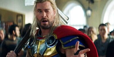 Thor, portrayed by chris hemsworth, looks shocked and disheveled with long blonde hair and a red cape, amidst a busy indoor scene.