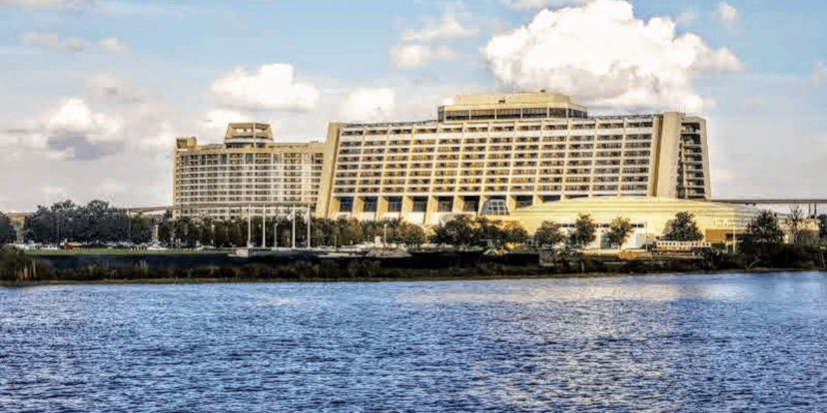 A large, multi-story hotel sits beside a body of water, with a landscaped foreground and a partly cloudy blue sky overhead.