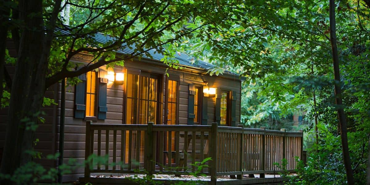 A cozy wooden cabin surrounded by lush green trees, with lights glowing warmly through its windows and illuminating the porch at dusk.