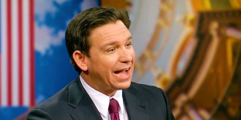 Governor Ron DeSantis speaking during a televised interview, with an american flag in the background.