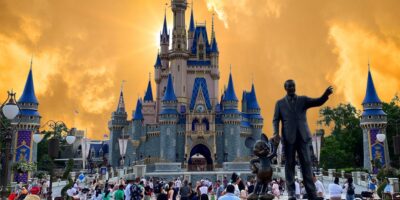 Sunset hues cast a dramatic backdrop over the iconic cinderella castle and the statue of walt disney and mickey mouse at a bustling disney theme park.