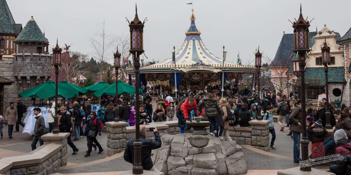 A lively day at a theme park with visitors milling around, a carousel in the background, and a stone well in the foreground.