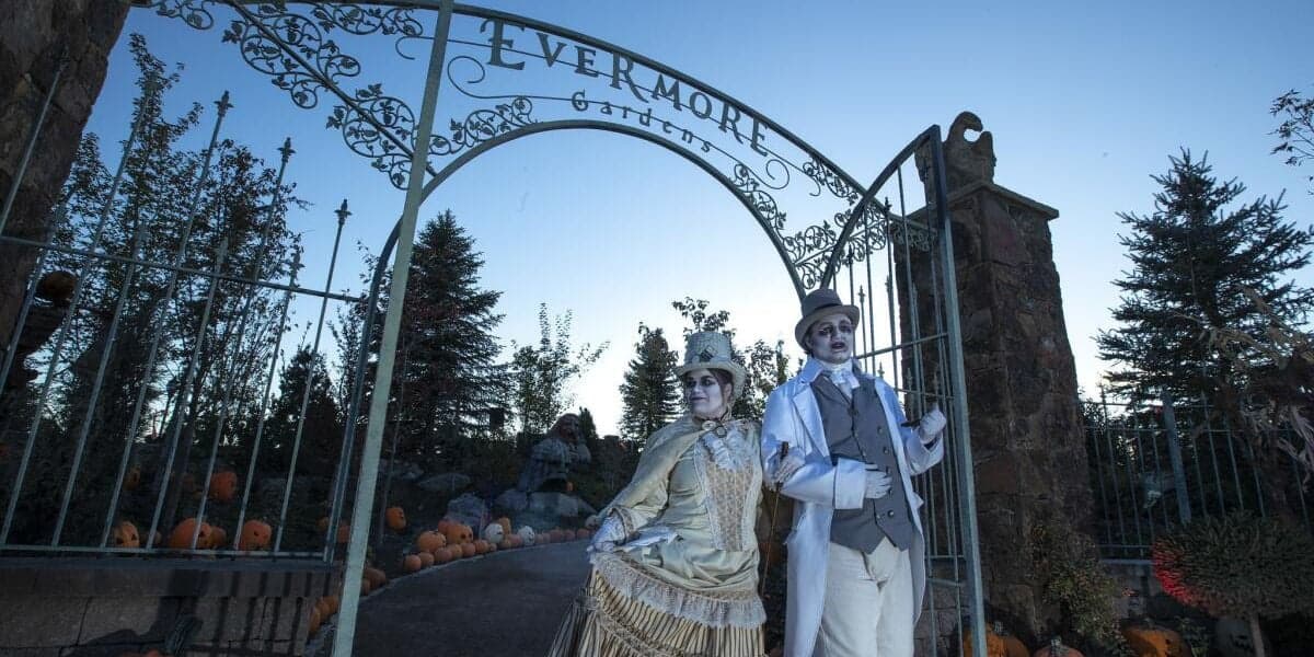 Two performers in vintage, whimsical costumes stand under an ornate metal arched gate labeled "evermore," surrounded by pumpkins and dusky sky.