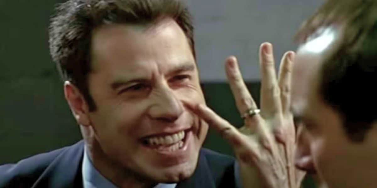 John Travolta smiling and waving hand in front of face in front of Nicolas Cage (right) in Face/Off.