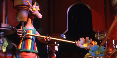A colorful animated dragon wearing a striped shirt holds a scepter, standing in a whimsical, mechanically decorated room with vibrant colors and arched doorways.
