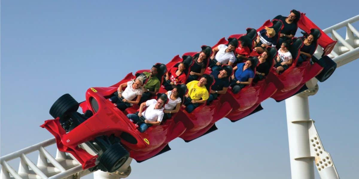 People experiencing the thrill of a roller coaster resembling a red sports car as they zoom down a steep track against a clear blue sky.
