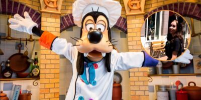 A Disney World character, Goofy, dressed as a chef stands with arms wide in a vibrant kitchen setting. In the corner, there's an inset photo of a woman sitting outdoors at a table, wearing a black outfit and sunglasses.
