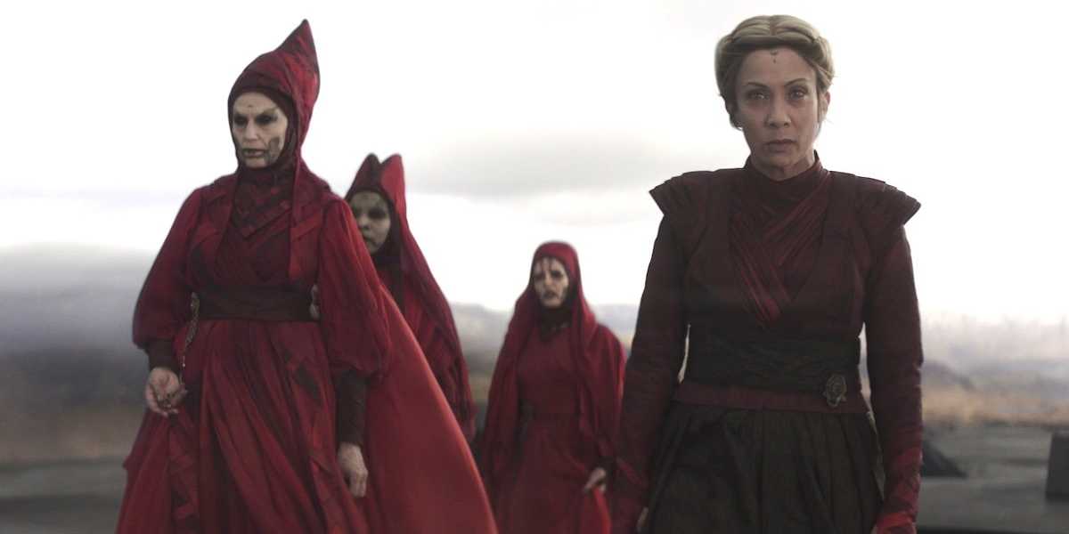 The Great Mothers and Morgan Elsbeth dressed in elaborate red costumes and headpieces walking solemnly in a dim, barren landscape. two figures in the foreground, one wearing a mask, the other unmasked with a stern expression.