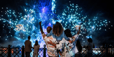 A group of people watches a vibrant fireworks display at night, with one person wearing glowing mickey mouse ears. they are standing by a railing, illuminated by the colorful bursts in the sky.