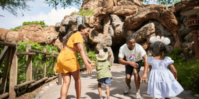 A family enjoying a sunny day outdoors; a woman in a yellow dress and a man in a striped shirt playfully chase two children wearing hats near an elaborate tree structure in a park.