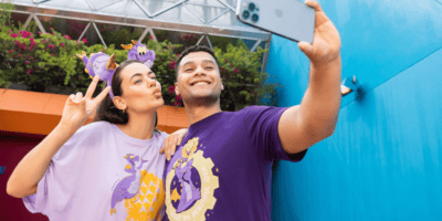 A young couple takes a selfie; the woman is making a peace sign. they wear matching purple t-shirts with cartoon dragon designs, standing against a vibrant blue backdrop.