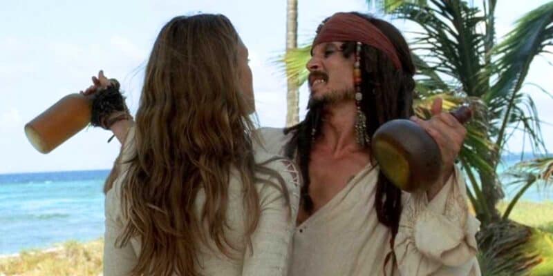 A man dressed like a pirate with long hair and bandana holds a bottle, standing next to a woman with long hair, both on a tropical beach.