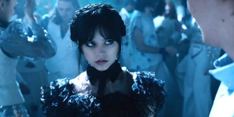 Jenna Ortega wearing a black ruffled dress stands amidst a dance floor, surrounded by mist and softly lit figures.