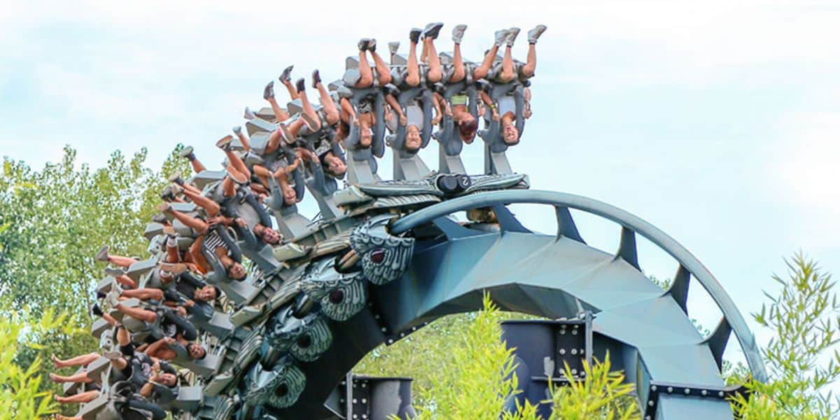 Roller coaster with inverted passengers plunging down a curved track amidst lush green trees under a blue sky.