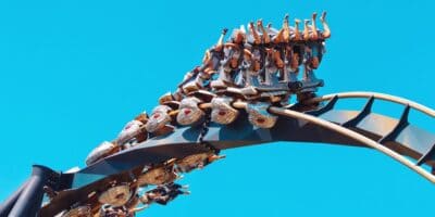 A roller coaster with passengers, arms raised, cresting a sharp turn against a clear blue sky.