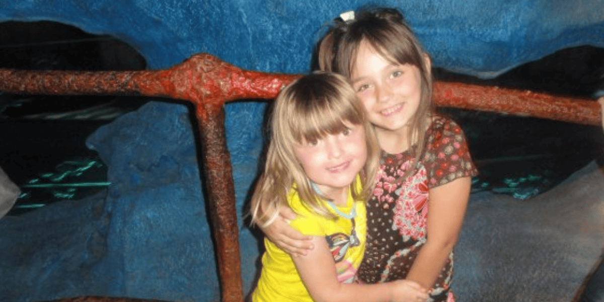 Two young girls with joyful expressions hugging each other in front of a blue, aquarium-like backdrop. one girl wears a yellow shirt, the other a floral top.