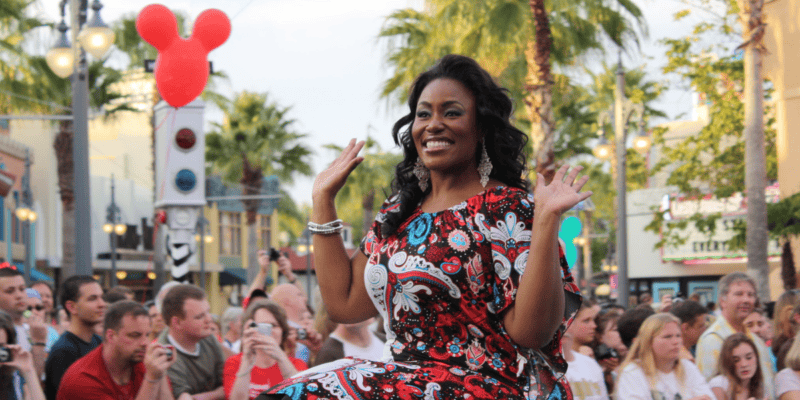 A joyful woman in a colorful patterned dress waves to a crowd during a parade, with large balloon and palm trees in the background.