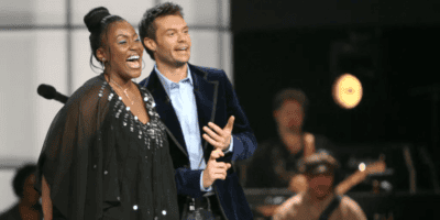 A woman and a man stand on a stage, singing and smiling together. the woman is wearing a sparkling black outfit, and the man is in a blue suit. a band plays in the background.