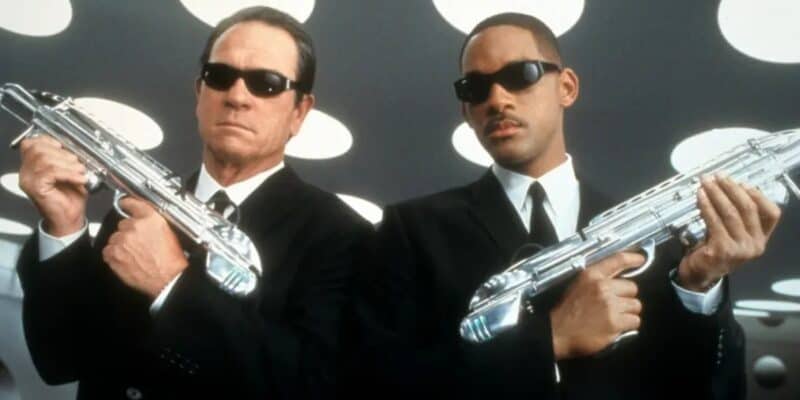 Tommy Lee Jones and Will Smith dressed in suits and sunglasses hold futuristic silver guns, standing in front of a wall with circular white patterns. they appear focused and ready for action.