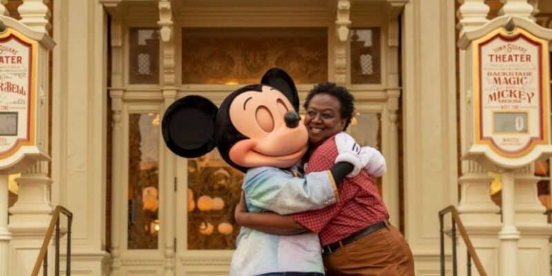 A joyful woman hugs mickey mouse in front of a theater with posters reading "backstage theater magic of mickey!" their expressions are cheerful.