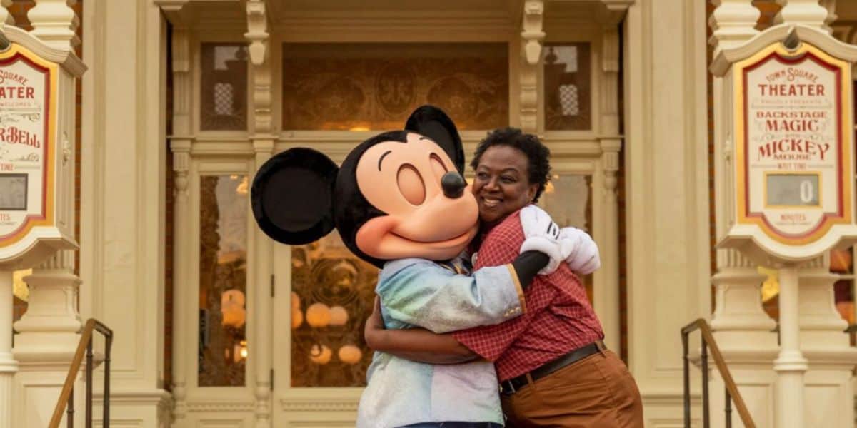 A joyful woman hugs mickey mouse in front of a theater with posters reading "backstage theater magic of mickey!" their expressions are cheerful.