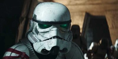 A stormtrooper with a battle-worn helmet and green-tinted goggles looks intently forward, standing in a dimly lit setting.