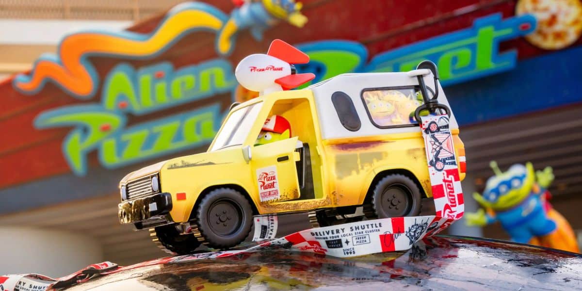 A vibrant toy delivery shuttle with decals, from the movie "toy story", parked atop a globe against a colorful backdrop featuring an "alien pizza planet" sign, for sale only at Pixar Fest.