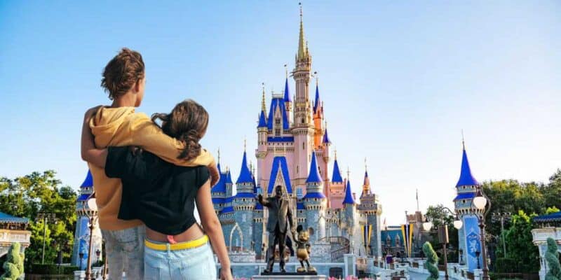 Two people from behind, one carrying the other piggyback, admiring the view of a fairy-tale castle under a clear blue sky.