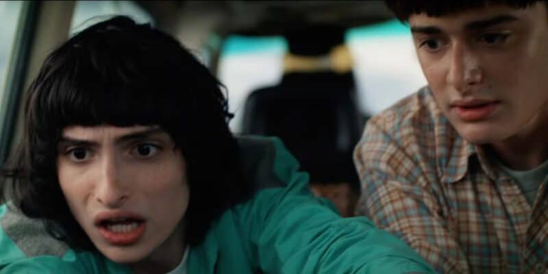 Two young adults, a woman with short black hair wearing a turquoise jacket and a man in a checkered shirt, look intensely out of a car window with anxious expressions.