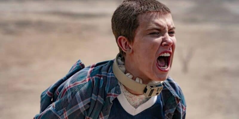 A young boy with a buzz cut and neck brace, wearing a plaid shirt, yells intensely in a barren, sandy landscape.