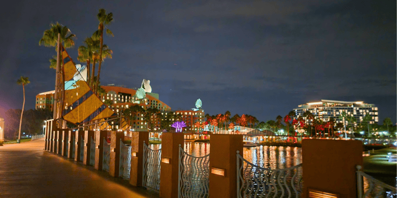 Night view of a waterfront promenade with illuminated figures and palm trees, a calm river reflecting city lights, and buildings in the background under a cloudy sky.
