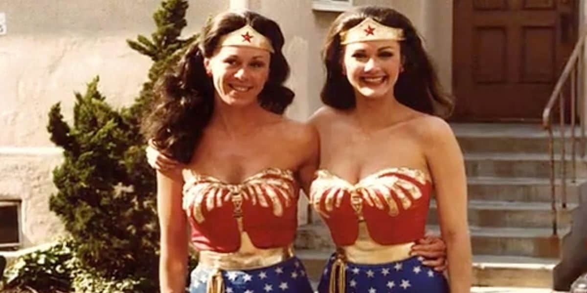 Two women dressed as Wonder Woman, standing side by side and smiling, in front of a building's staircase. They wear matching costumes with iconic red, blue, and gold detailing.