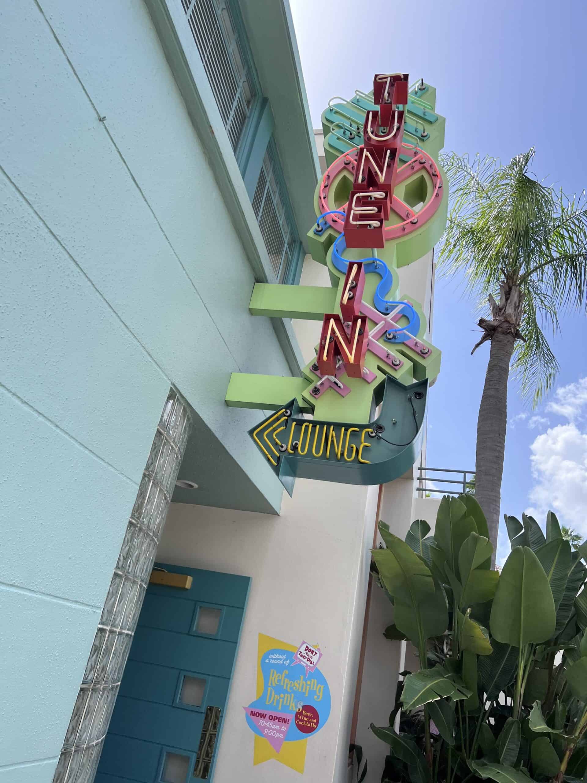 Colorful neon sign that reads "Tune-In Lounge" in vibrant pink and green, attached to a light blue building under a clear blue sky, with a palm tree in the background at Disney World.