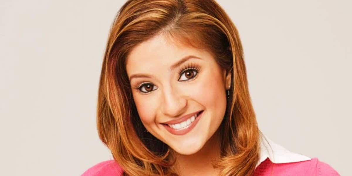 Anneliese van der Pol with long, light brown hair and large eyes smiling at the camera, wearing a pink top with a white collar.