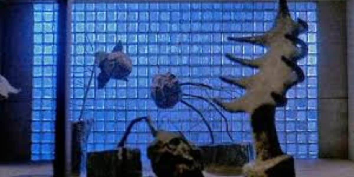 A dimly lit room features abstract sculptures coated with snow, reminiscent of Tim Burton's distinct style in movies like Beetlejuice. A slender tree-shaped figure and several spherical objects with tendrils adorn the scene, while the background is illuminated by a blue-tinted grid of frosted glass blocks.