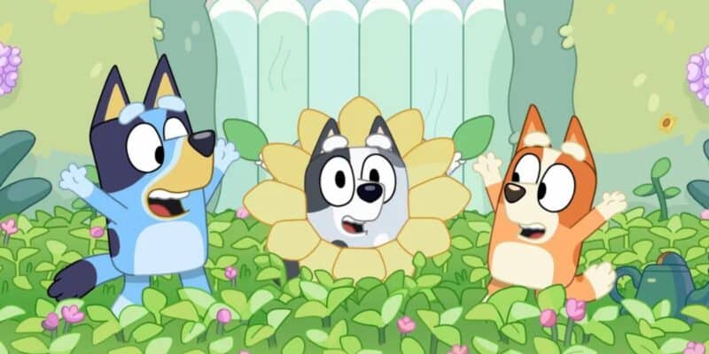 Three animated dog characters are playing in a lush garden. The blue dog on the left and the orange dog on the right are enthusiastically raising their arms. The central character, with a gray face, is dressed as a flower. They are surrounded by greenery, flowers, and even some muffin-shaped bushes.