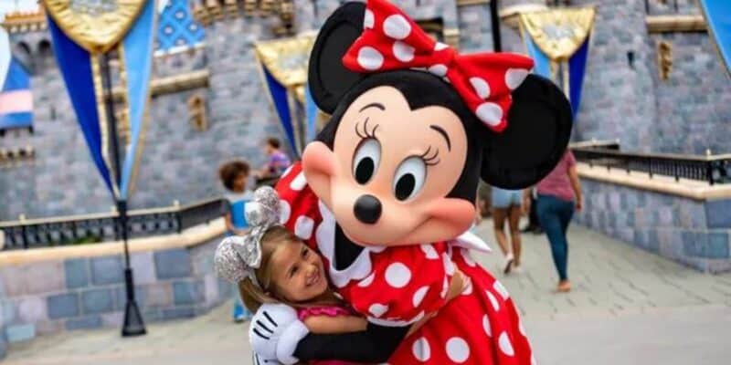 A child wearing silver Mickey Mouse ears hugs a person in a Minnie Mouse costume at Disneyland, where character actors recently unionized. The castle and other visitors are visible in the background, and both the child and the character appear to be happy.