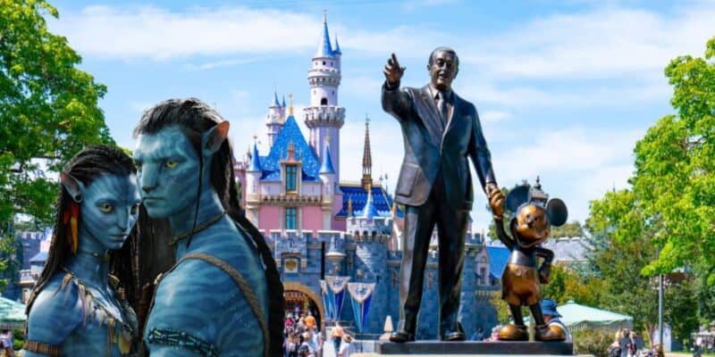 Two characters from the movie "Avatar" are digitally added in front of the Sleeping Beauty Castle at Disneyland. They stand beside the "Partners" statue of Walt Disney holding hands with Mickey Mouse, blending something new with fantasy elements and the iconic theme park backdrop.