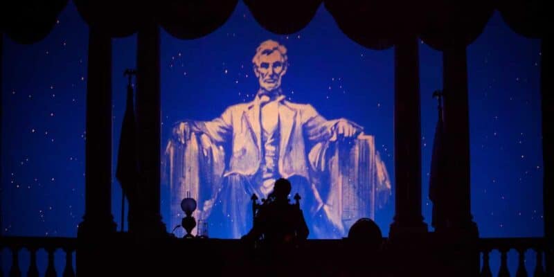 Silhouettes of people watch an illuminated projection of Abraham Lincoln sitting in a chair at the "Great Moments With Mr. Lincoln" exhibit, framed by a decorative arch and a starry background.