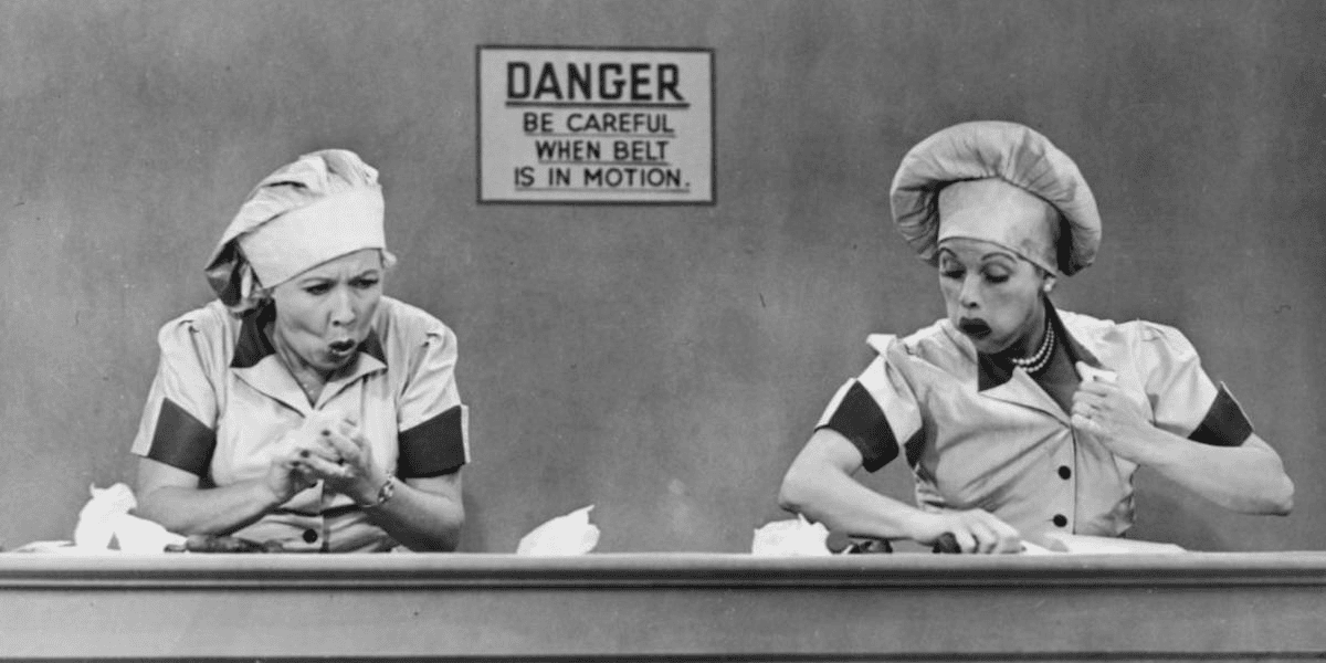 Two women wearing chef hats and uniforms frantically work at a conveyor belt with a "DANGER" sign behind them, reminiscent of Lucille Ball's comedic chaos. Overwhelmed as they handle items on the belt, the black-and-white scene captures the spirit of vintage television hilarity.