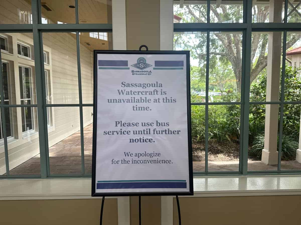 Sign on an easel stating "Disney sassagoula watercraft is unavailable at this time. Please use bus service until further notice." located inside a building with windows looking out to trees.