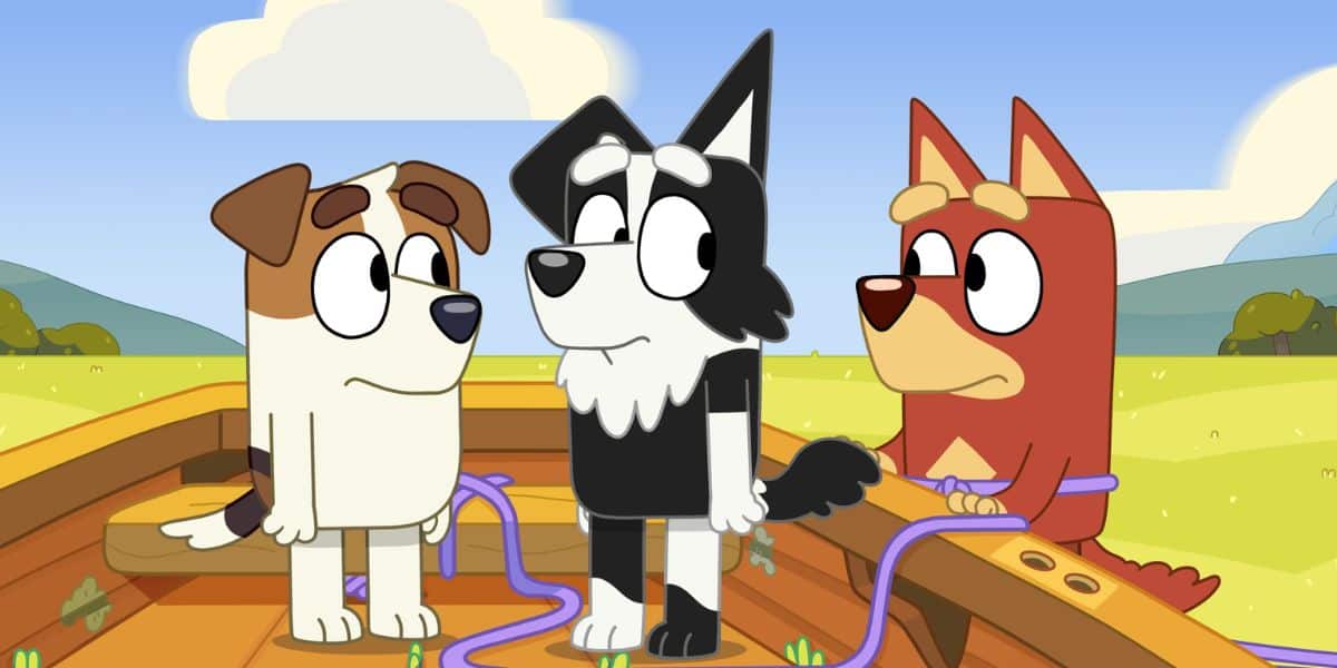 Three cartoon dogs, a brown and white dog named Bluey, a black and white dog, and a red dog, stand on a wooden platform in a grassy field with a clear sky.