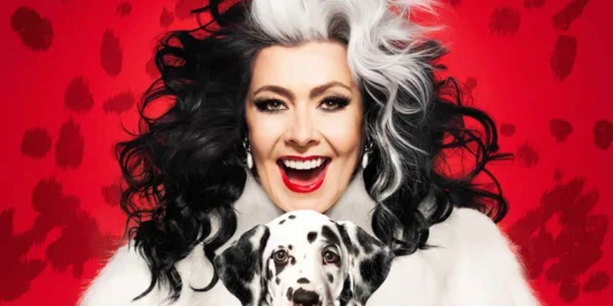 A woman with striking black and white hair, bright red lipstick, and a joyful expression holds a Dalmatian puppy. The background is red with black spots, reminiscent of 101 Dalmatians. She is wearing a white fur coat.