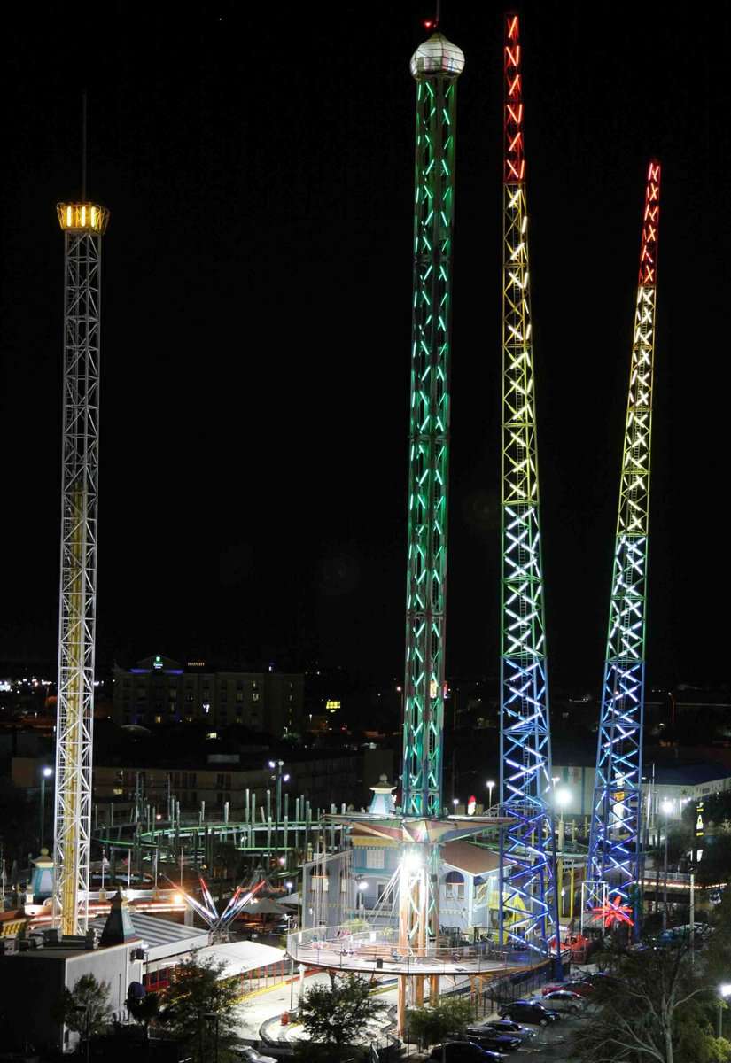 Three brightly lit amusement park towers at night inside Magical Midway Thrill Park, each glowing in different colored lights, with surrounding attractions and buildings.