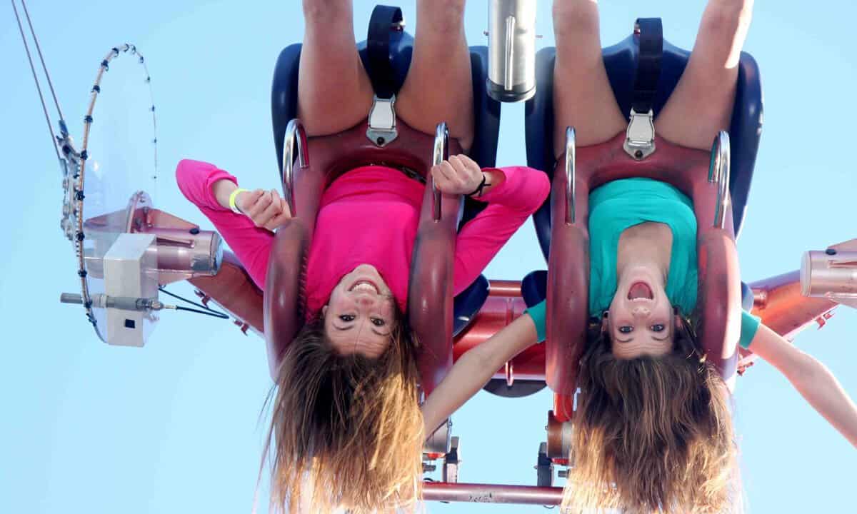 Two girls with long hair hanging upside down and laughing on a ride inside of Midway Magical Thrill Park, dressed in bright pink and green tops against a clear blue sky.