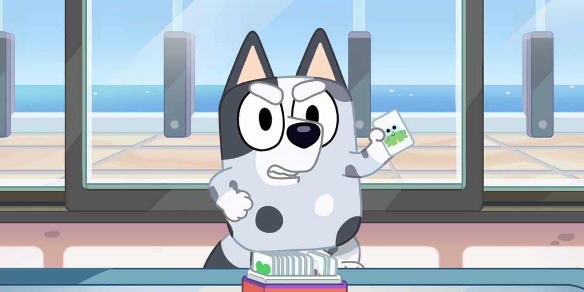 Animated scene featuring a cartoon dog with grey and white fur standing behind a counter. The dog has a stern expression and is holding up a piece of paper with a green logo, while next to him sits an untouched muffin. There's a large window in the background showing an ocean view.