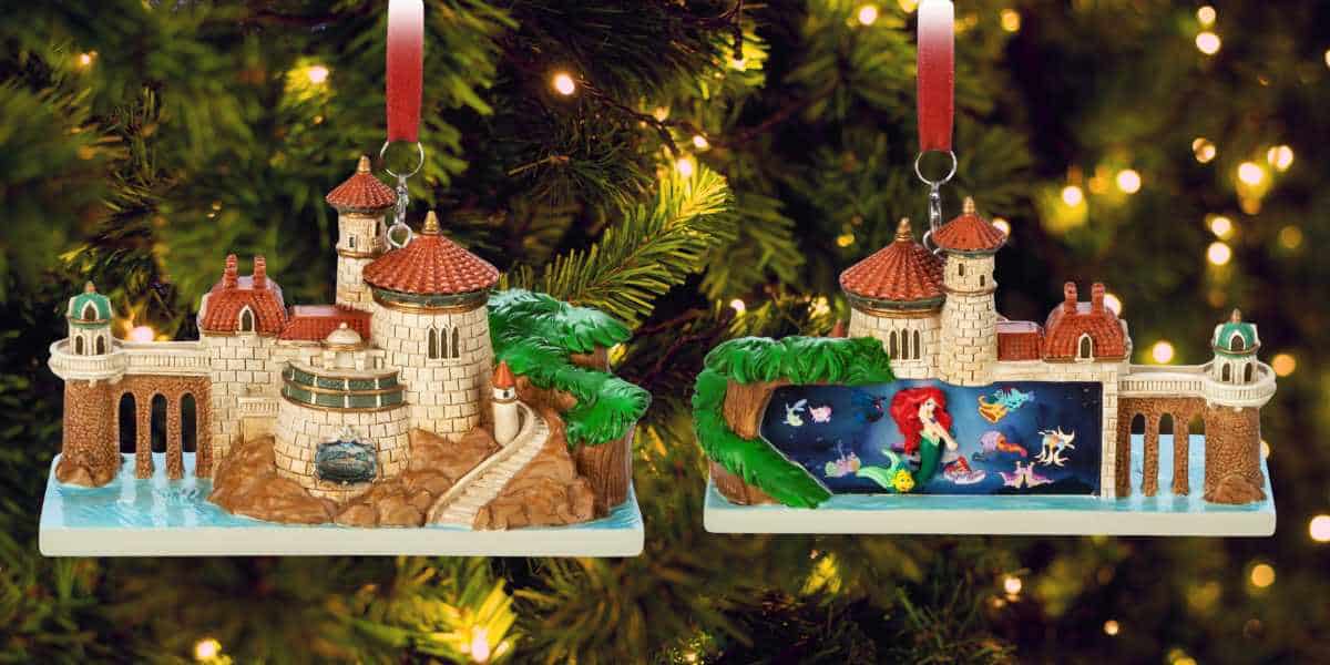 Two Disney park-themed ornaments depicting scenes from "The Little Mermaid" hang on a Christmas tree. The left ornament shows an elaborate castle, while the right ornament features Ariel and her underwater friends in a vibrant aquatic scene. The tree is adorned with lights, adding to its festive charm.